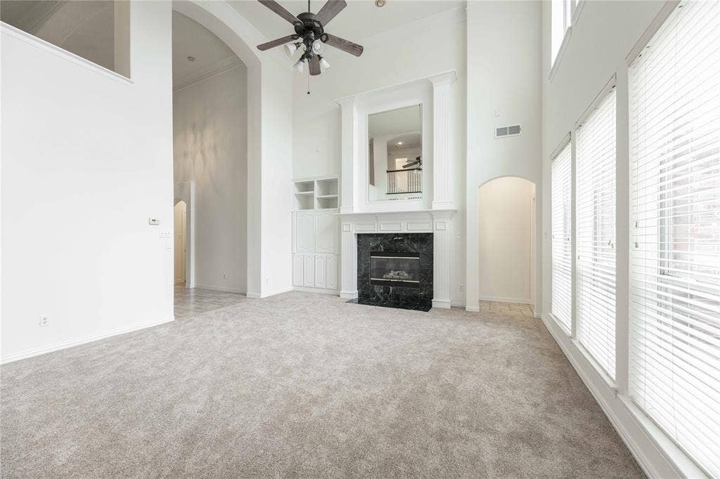 3305 Estacado Lane, Plano, TX 75025 Spacious living area featuring high ceilings, a fireplace with a dark stone surround, tall windows with blinds, a ceiling fan, and light-toned carpeting