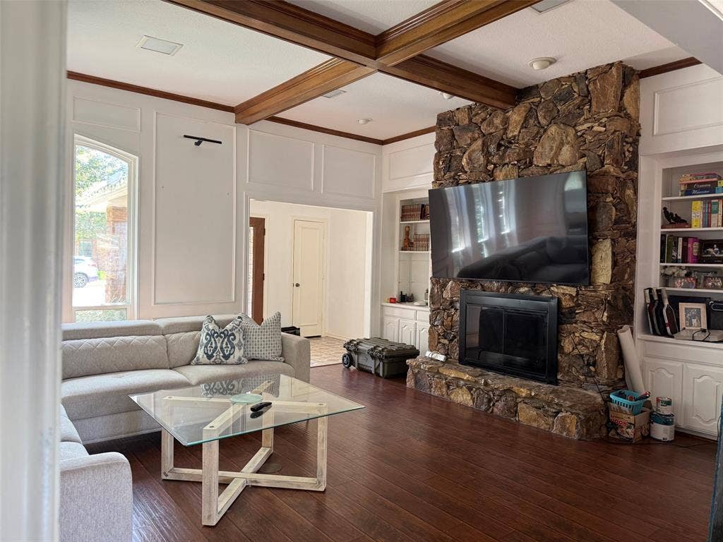 1517 Aylesbury Lane, Plano, TX 75075 Stone-clad fireplace with hearth, built-in shelving, wood-finish flooring, exposed wood ceiling beams, and arched window