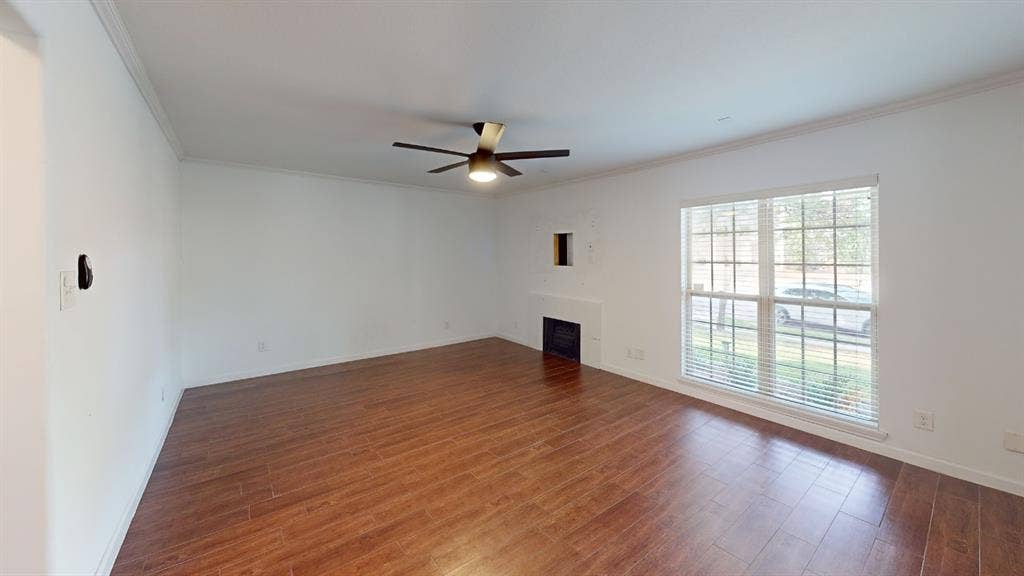 Wood-finish flooring, ceiling fan, white crown molding, fireplace with mantel, and large window with blinds