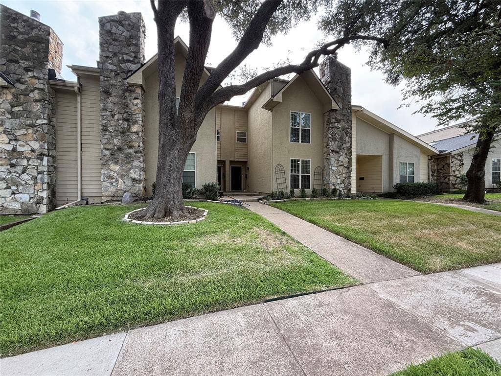 Exterior featuring stone veneer accents, stucco siding, a mature tree, an attached covered entryway, and a concrete walkway