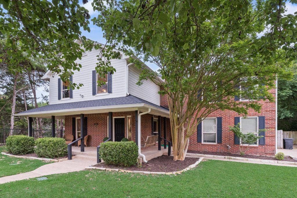 Welcoming exterior featuring a classic brick-and-siding design, mature shade trees, manicured landscaping, and a spacious covered front porch.
