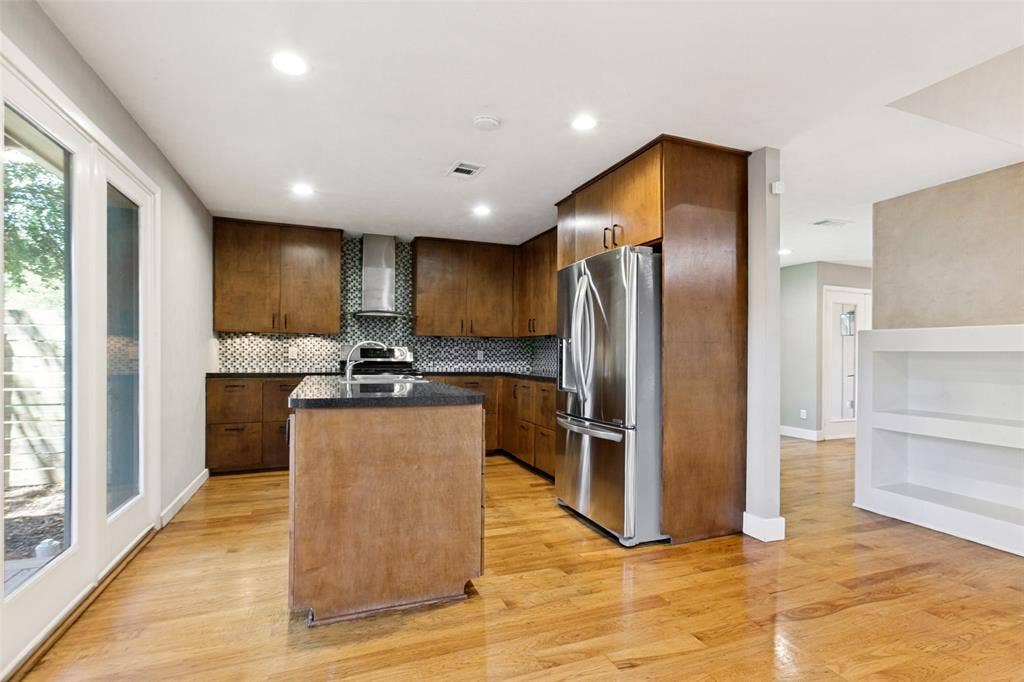 2937 Forest Hills Lane, Richardson, TX 75080 Kitchen featuring wood cabinetry, stainless steel appliances, a center island with a dark countertop, a patterned tile backsplash, and wood-finish flooring