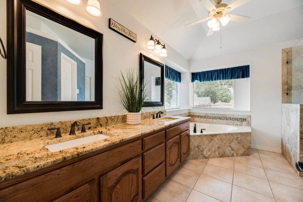 1485 Ashbourne Drive, Rockwall, TX 75087 Dual vanity with granite countertops, wood cabinetry, built-in soaking tub with tile surround, separate tiled shower, and vaulted ceiling with ceiling fan