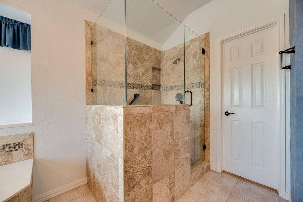 1485 Ashbourne Drive, Rockwall, TX 75087 Tiled shower enclosure with a clear glass door, built-in shelving, and a decorative tile accent band