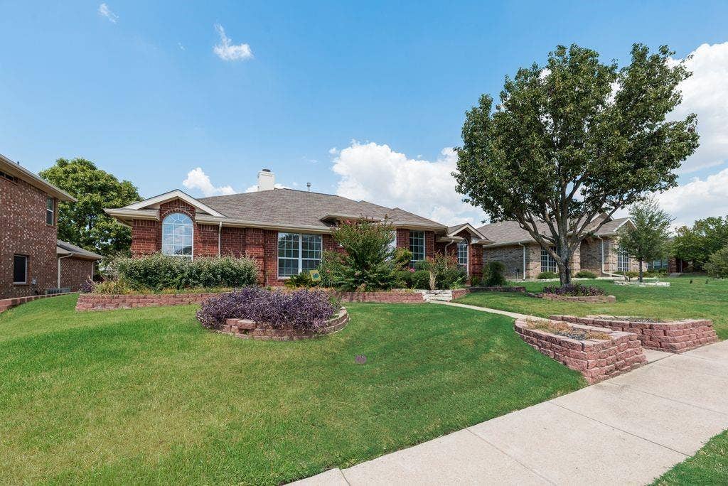 1485 Ashbourne Drive, Rockwall, TX 75087 Brick exterior with arched window, concrete walkway, mature trees, and landscaped garden beds