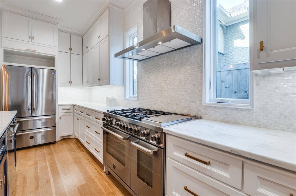 4031 Prescott Avenue, Dallas, TX 75219 White cabinetry with gold-tone hardware, stainless steel appliances, wood-finish flooring, and a white tile backsplash