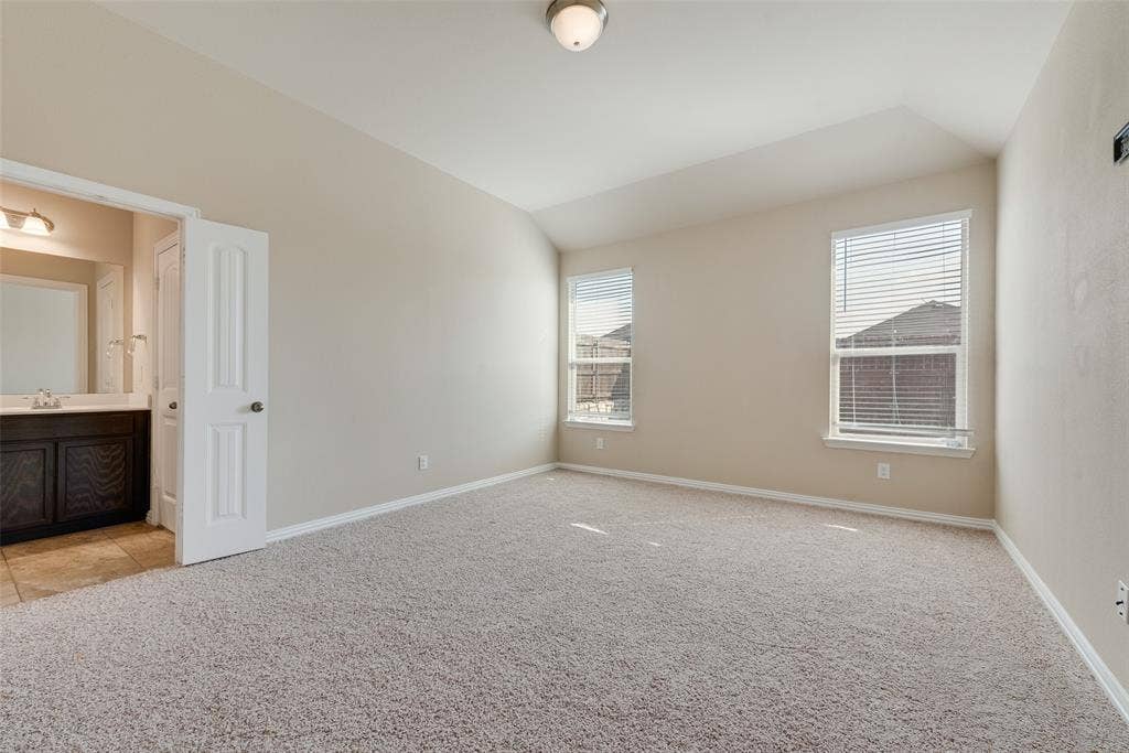 215 Fred Lane, Ferris, TX 75125 Vaulted ceiling, two windows with blinds, neutral carpeting, light neutral wall paint, and an en-suite bathroom featuring a vanity with a mirror and lighting