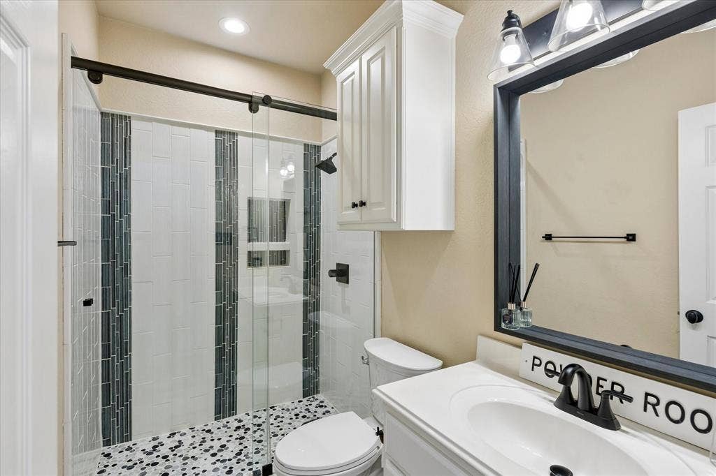 3800 Deer Forest Drive, Denton, TX 76208 Glass-enclosed shower with subway tile and mosaic accents, pebble-style shower floor, white vanity with integrated sink, framed vanity mirror, and overhead cabinet storage