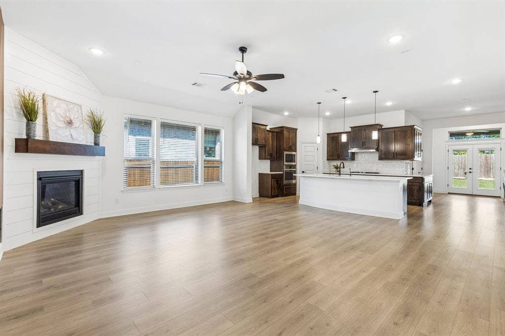 5224 Troupe Road, McKinney, TX 75071 Spacious room featuring wood-finish flooring, a fireplace with a wood mantel, a kitchen island with pendant lighting, dark wood cabinetry, and double glass-paneled doors