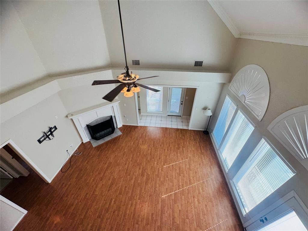 Vaulted ceiling, wood-finish flooring, white mantel fireplace, ceiling fan with integrated lighting, and multiple windows with blinds