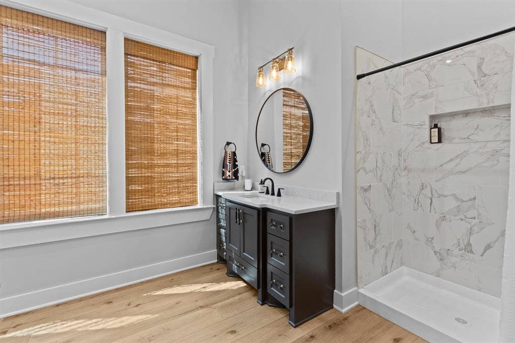 23180 Bridle View Drive, Lindale, TX 75771 Wood-finish flooring, dark wood vanity with white countertop, round wall-mounted mirror, walk-in shower with tile surround, and dual windows with natural woven shades