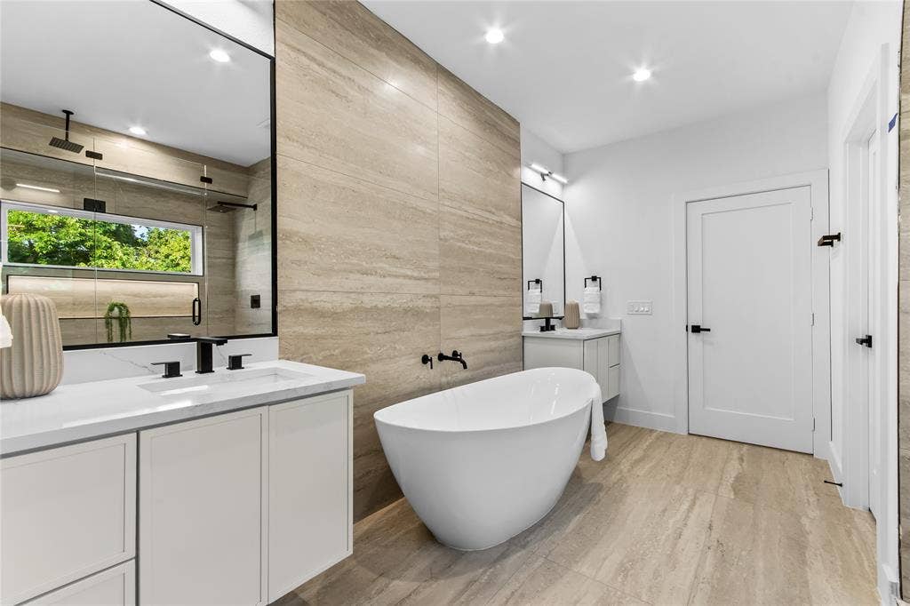 Contemporary bathroom featuring a freestanding soaking tub, dual vanities with white cabinetry and integrated sinks, a walk-in shower with a rain shower head, and floor-to-ceiling textured tile accents