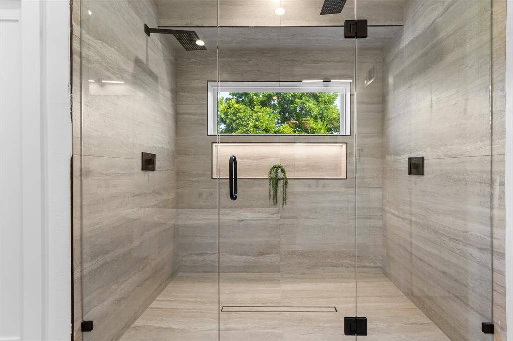 Frameless glass shower enclosure featuring large format stone-look tile, dual rainfall showerheads, an illuminated niche, and a linear drain