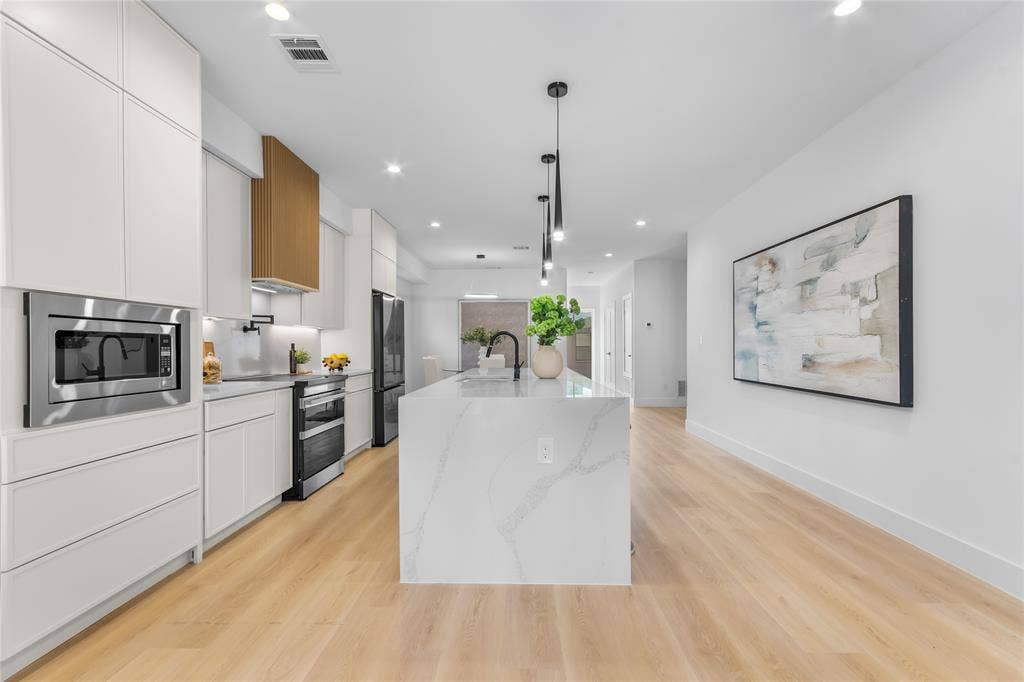 Contemporary kitchen featuring a spacious island with a waterfall-edge countertop, light wood-finish flooring, white flat-panel cabinetry, a stainless steel built-in microwave, and pendant lighting