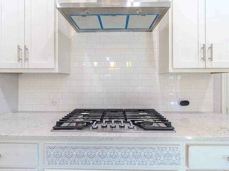Stainless steel gas range with a coordinating hood, white subway tile backsplash, white shaker-style cabinetry, and light-toned countertops