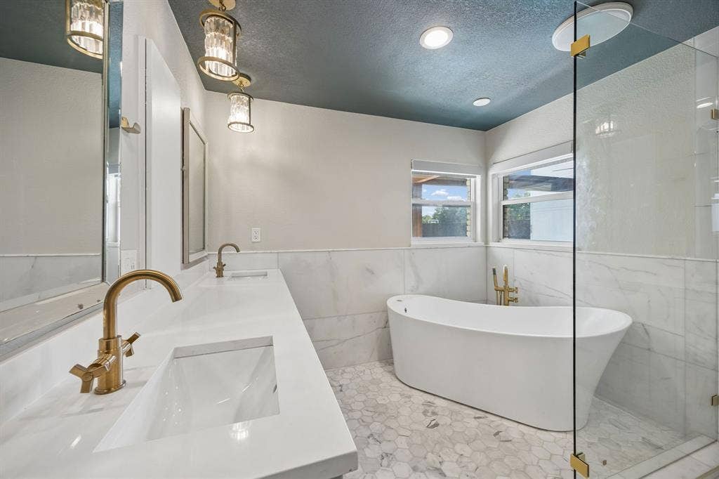 Beautifully updated primary bath featuring marble finishes, dual vanities, a freestanding soaking tub, glass-enclosed shower and elegant gold fixtures.