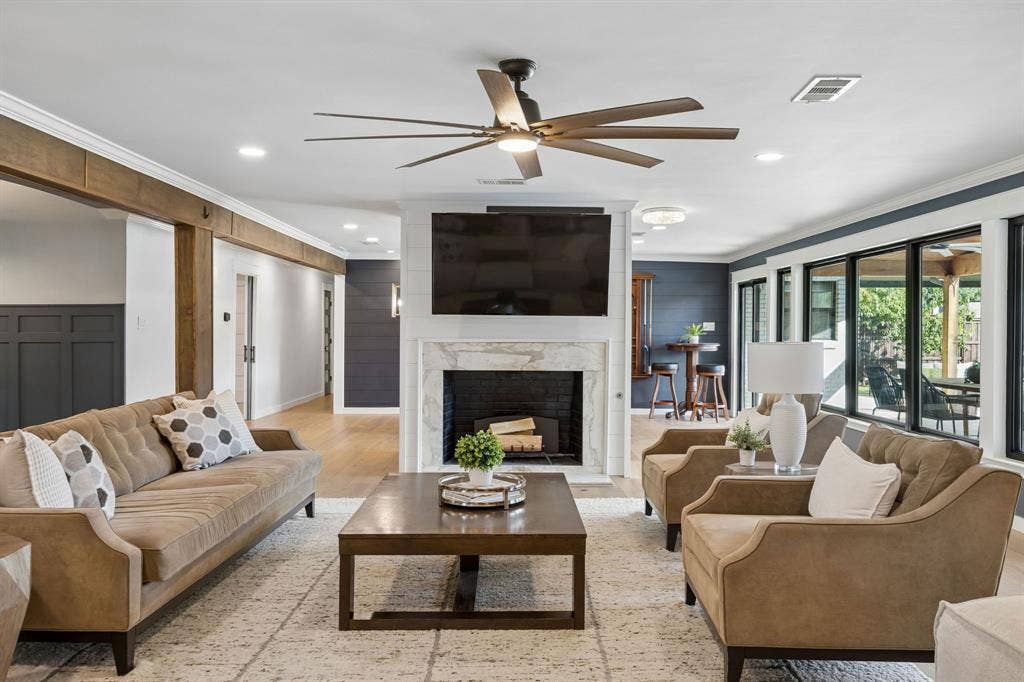 Inviting main living room featuring a wood burning gas fireplace with marble surround, wood floors, decorative wall treatments and a wall of windows overlooking the backyard.