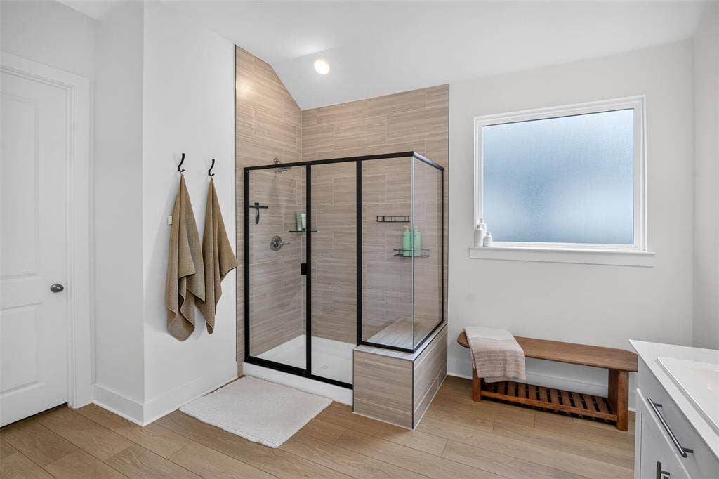 Spacious bathroom featuring a glass-enclosed shower with tiled walls and a built-in bench, wood-finish flooring, and a window with frosted glass