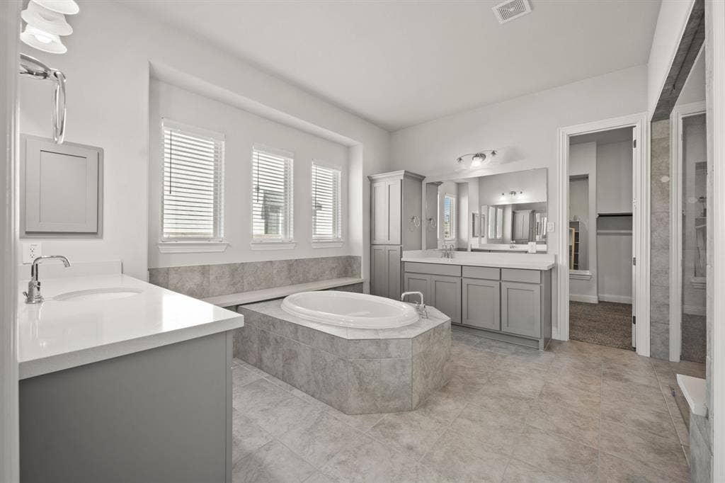 Dual vanity with undermount sinks and light countertops, a built-in soaking tub with tile surround, tile flooring, and multiple windows with blinds