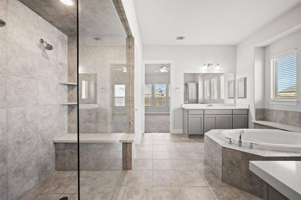 Spacious bathroom featuring a glass-enclosed shower with built-in bench and shelving, a separate soaking tub, a double vanity with grey cabinetry, and large format tile flooring