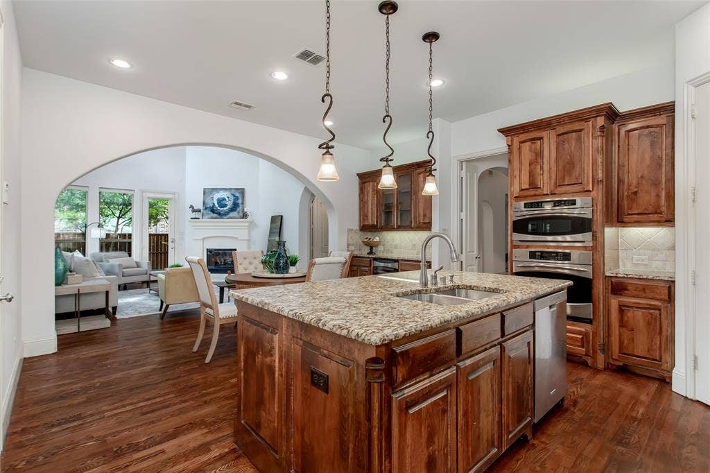 Gourmet kitchen featuring a central island with a double basin sink and granite countertop, rich wood-finish cabinetry, stainless steel double ovens, and recessed lighting