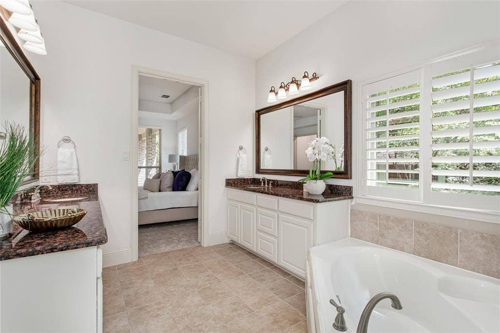 Dual vanity with dark stone countertops, white cabinetry, a large framed mirror, and an adjacent soaking tub