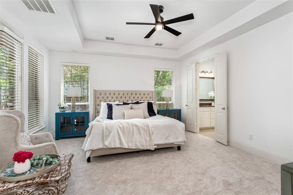 Tray ceiling with a ceiling fan, light-colored carpeting, multiple windows with blinds, and white double doors leading to a bathroom