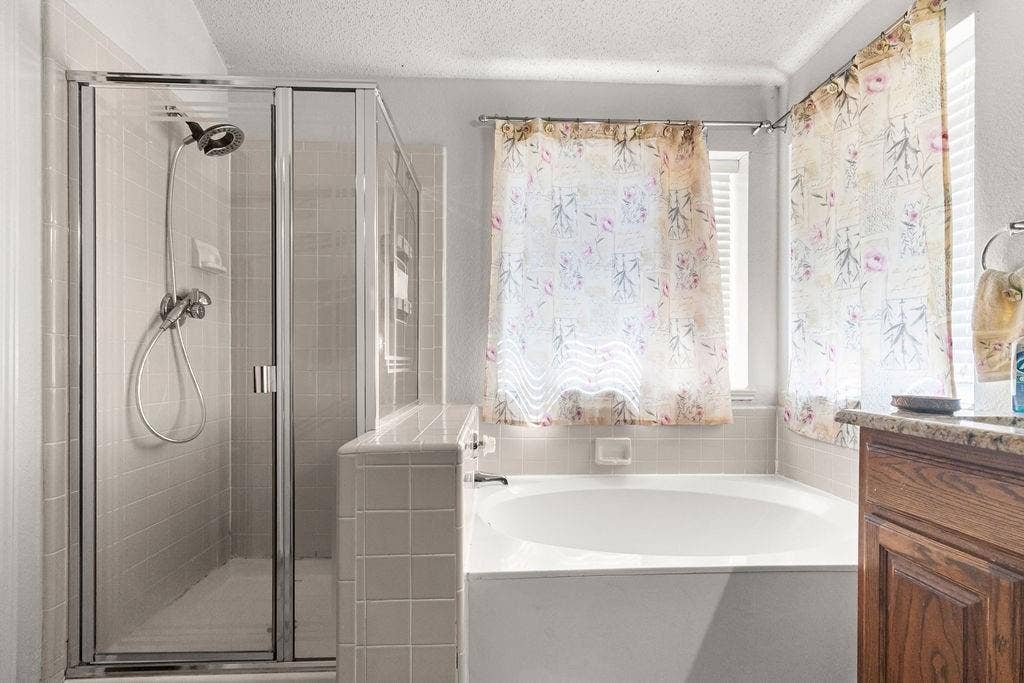 926 Wintercrest Court, Arlington, TX 76017 Tiled shower enclosure with a glass door, built-in bathtub, and a wood-finish vanity with a stone-finish countertop