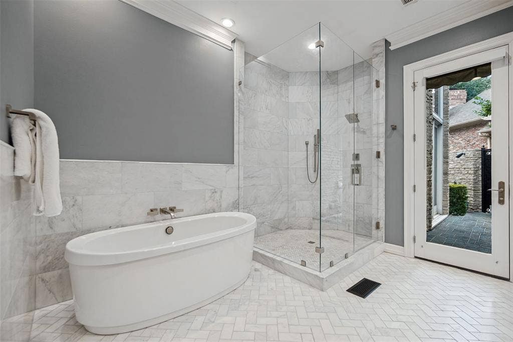 Freestanding soaking tub, walk-in shower with clear glass enclosure, marble-style tile walls, herringbone tile flooring, and direct outdoor access