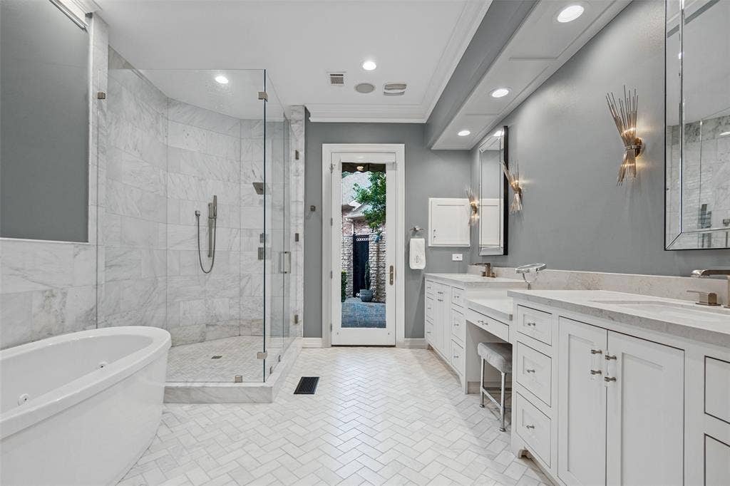 Freestanding soaking tub, walk-in shower with glass enclosure, dual vanity with undermount sinks, white cabinetry, and herringbone tile flooring