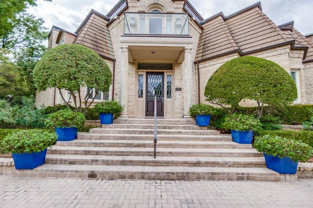 Light-toned brick exterior with a prominent entrance featuring a dark wood door, stone steps, and a glass-paneled balcony