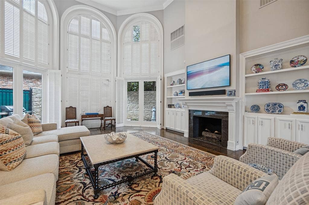 Two-story arched windows with plantation shutters, dark wood-finish flooring, built-in shelving, white fireplace surround, and a dark stone fireplace hearth