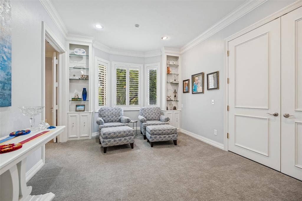 Built-in shelving with glass fronts, bay windows with plantation shutters, crown molding, recessed lighting, and carpet flooring