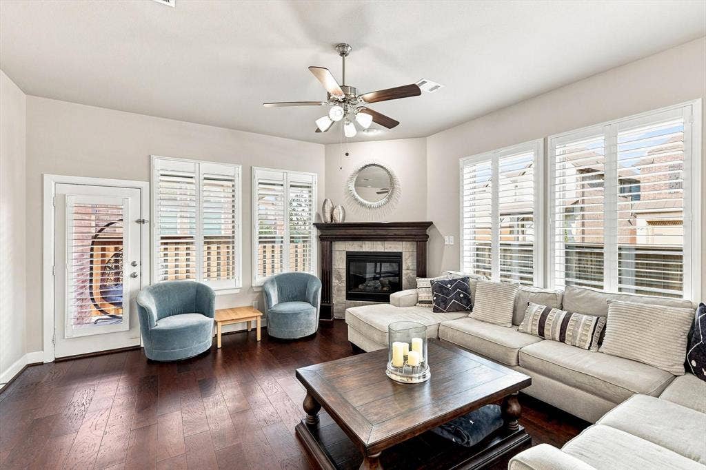 5441 Caine Road, Richardson, TX 75082 Hardwood flooring throughout, multiple windows with plantation shutters, ceiling fan with integrated lighting, and a corner fireplace with a dark wood mantel and tile surround