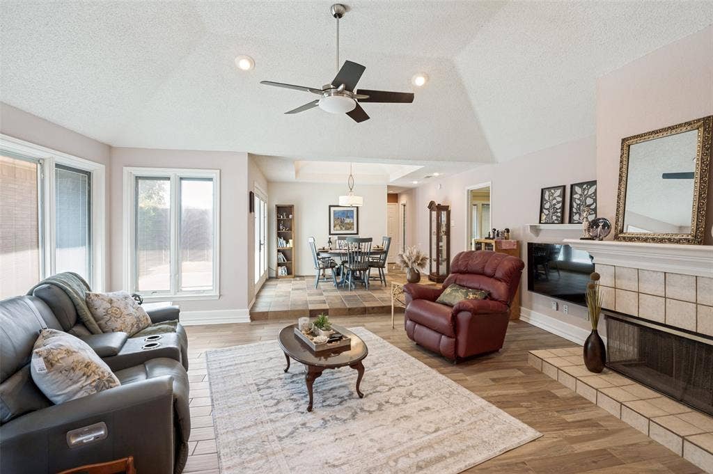 Vaulted ceiling with recessed lighting and ceiling fan, wood-finish flooring, tiled fireplace with mantel, and multiple large windows