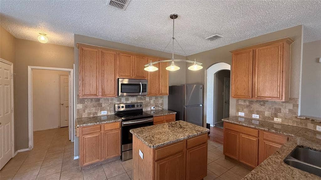 1433 Winterwood Drive, Allen, TX 75002 Kitchen featuring an island, light wood-finish cabinetry, stone countertops, stainless steel appliances, and a tile backsplash