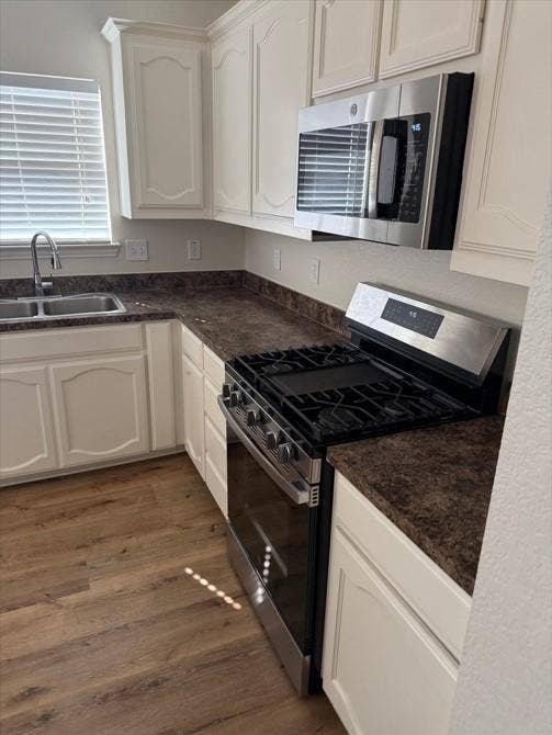 White shaker-style cabinetry with crown molding, stainless steel sink with gooseneck faucet, dark speckled countertops, stainless steel gas range, and wood-finish flooring