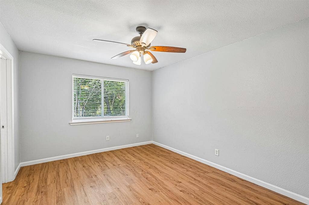 Bright secondary bedroom showcasing wood-finish flooring, white walls, and abundant natural light from a large double-hung window
