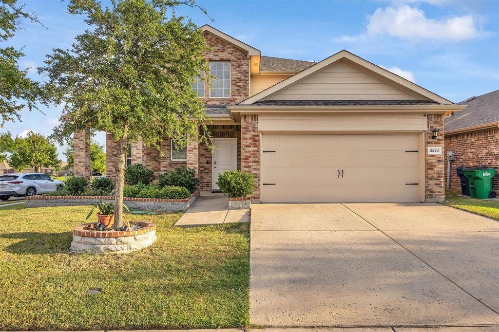 Brick facade with stone accents, two-story construction, attached garage, concrete driveway, and landscaped front yard with a stone-lined tree planter