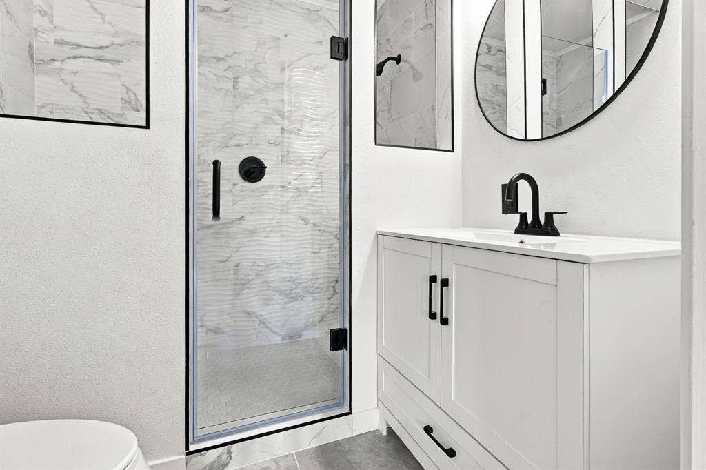 Walk-in shower with glass enclosure, single vanity with white countertop, matte black fixtures, and a frameless oval mirror