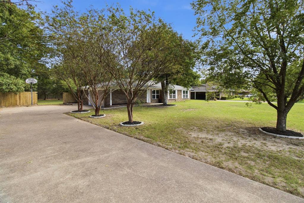 Expansive concrete driveway, mature trees, manicured front lawn, and wood privacy fencing