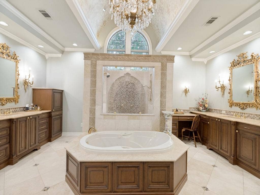 Vaulted ceiling with an ornate chandelier, a built-in jetted tub with stone surround, dual vanities with wood-finish cabinetry, and a walk-in shower with mosaic tile