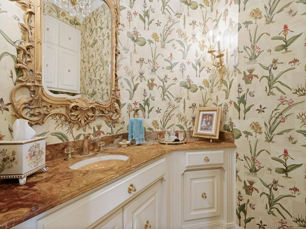 Ornate gold-framed mirror, botanical-patterned wallpaper, built-in cabinetry, polished stone countertop, and brass fixtures