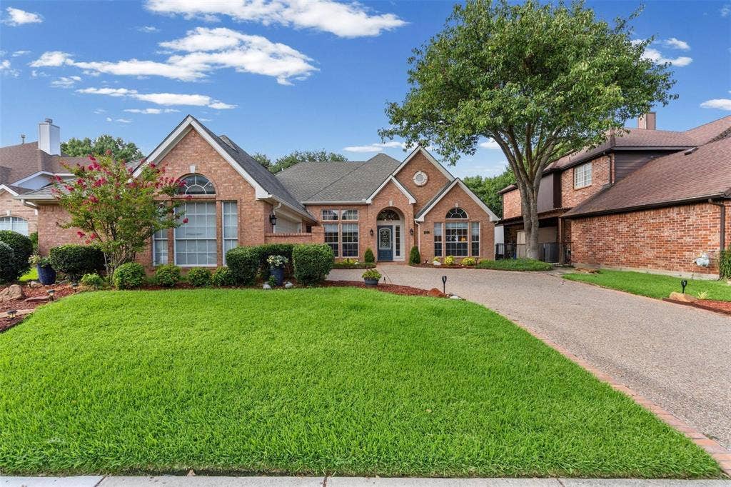 521 Palm Desert Drive, Garland, TX 75044 Brick Colonial home featuring a manicured front lawn, curved driveway, arched entryway with transom window, and mature landscaping including flowering shrubs and established trees