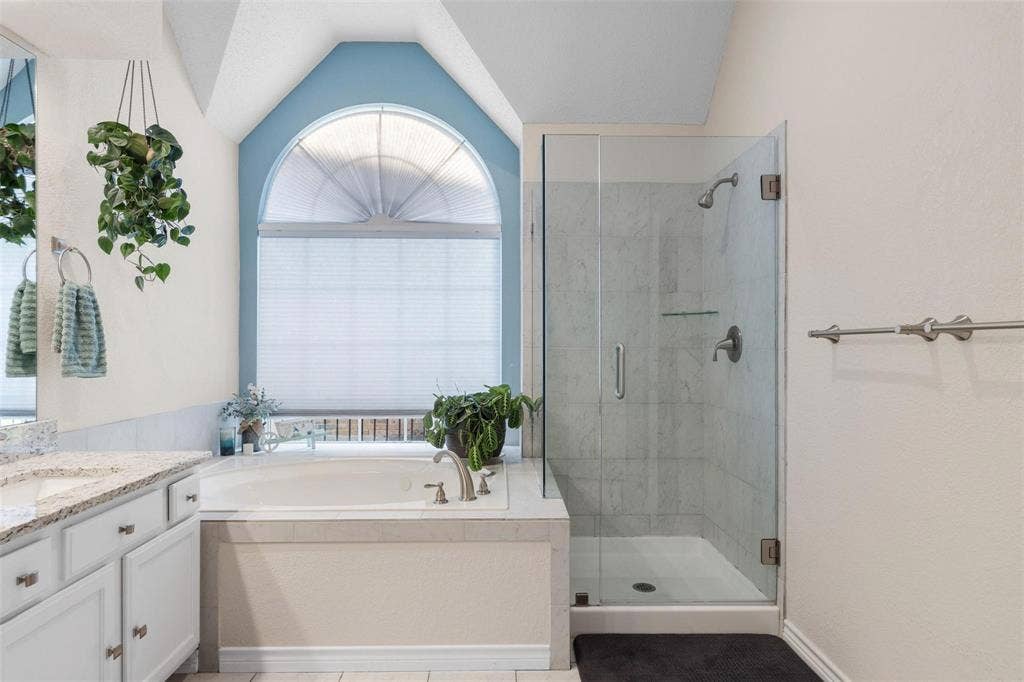 521 Palm Desert Drive, Garland, TX 75044 Built-in soaking tub, arched window with privacy blinds, glass-enclosed shower, white vanity with granite-style countertop, and tile flooring
