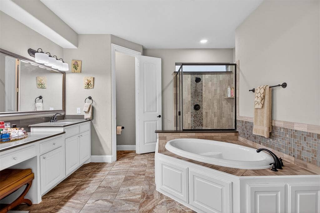 Spacious bathroom featuring a built-in soaking tub, glass-enclosed shower with tile surround, dual vanity with white cabinetry, and wood-finish flooring