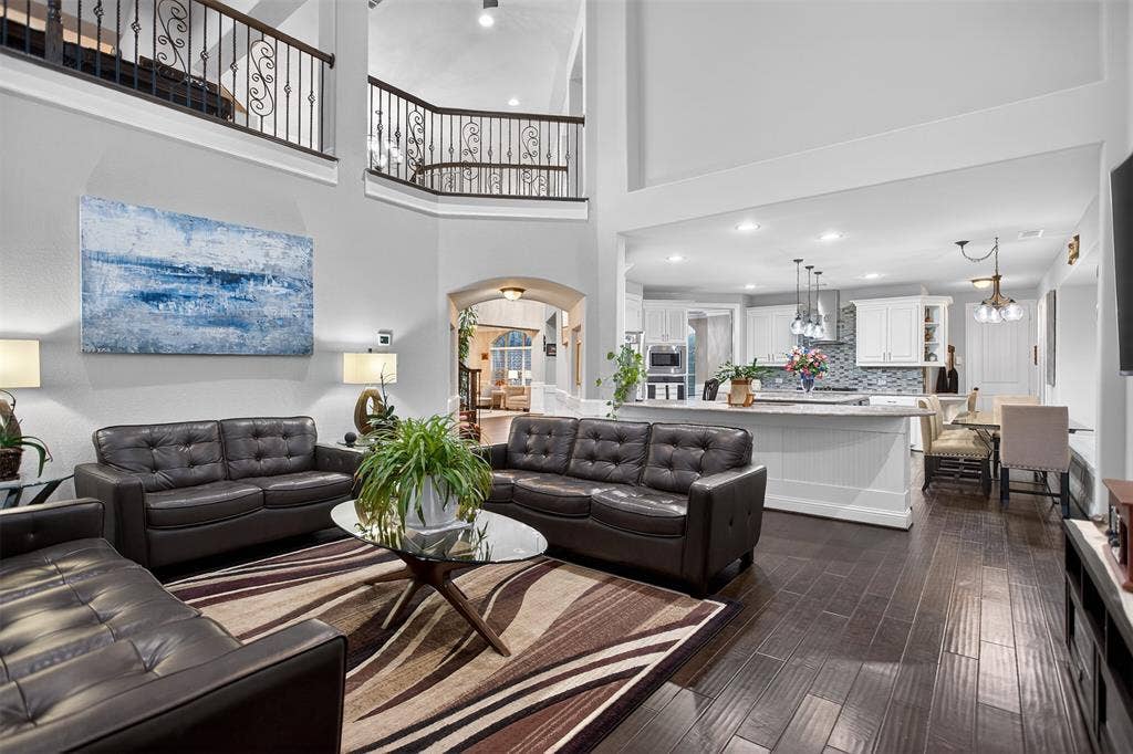 Two-story living area with dark wood-finish flooring, wrought iron railings, and a kitchen featuring white cabinetry, pendant lighting, and a tile backsplash