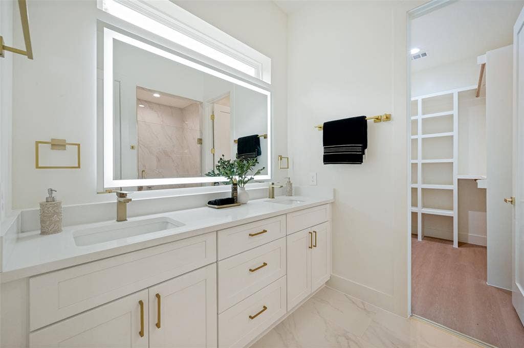 1511 Morrell Avenue, Dallas, TX 75203 Dual vanity with white cabinetry and gold-finish hardware, illuminated mirror, and a walk-in closet with built-in shelving