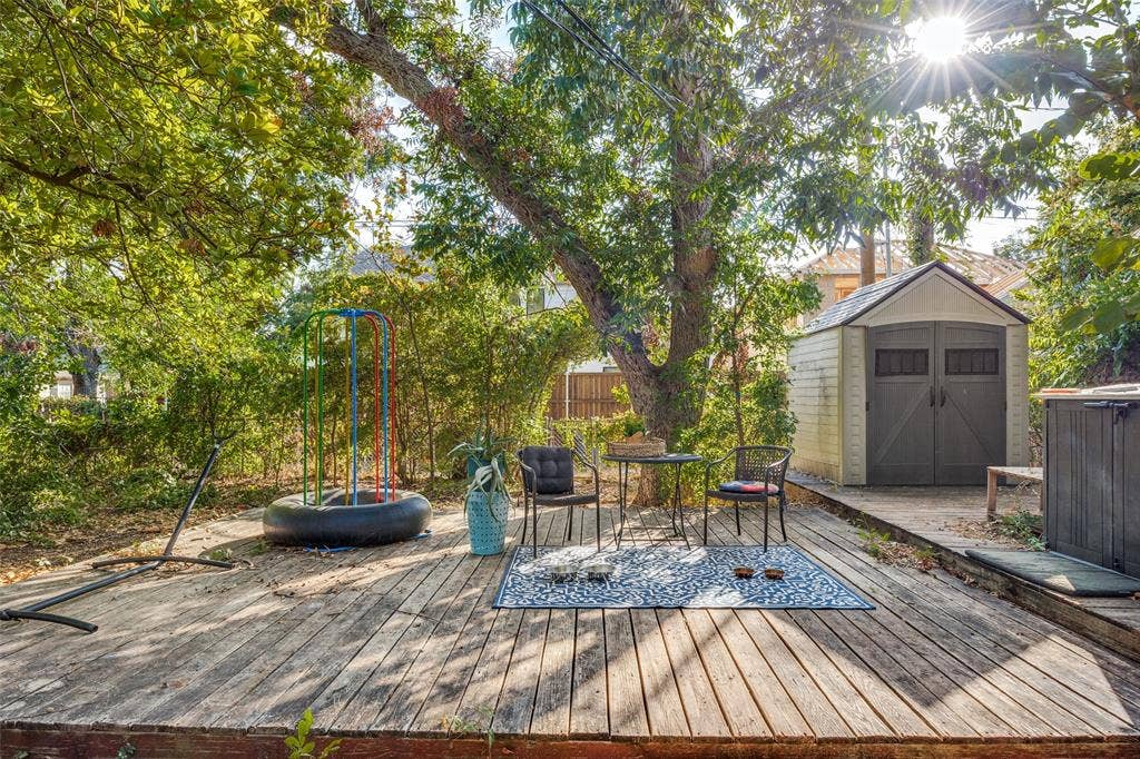 559 Blanning Drive, Dallas, TX 75218 Wood-deck patio area featuring 2 storage sheds with double doors and a pitched roof, mature trees, and dense perimeter foliage