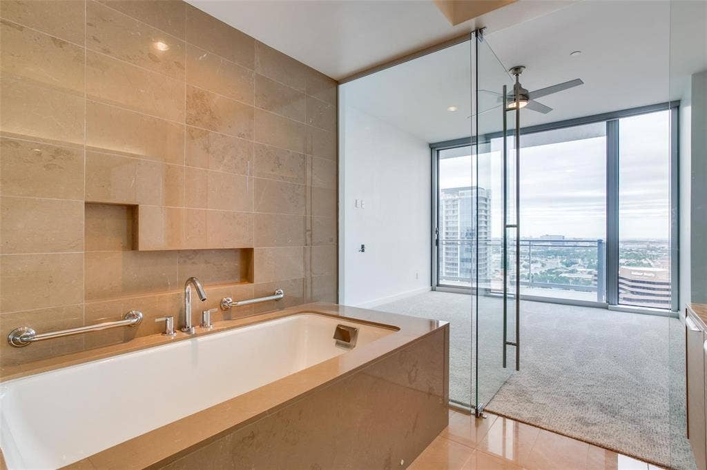Soaking tub with integrated shelf and chrome fixtures, full-height tile surround, glass partition, floor-to-ceiling windows, and ceiling fan